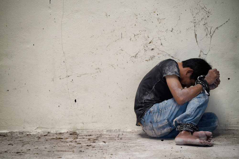 A barefoot person looking away from the camera, tied up with rope, sitting on a dirty floor.