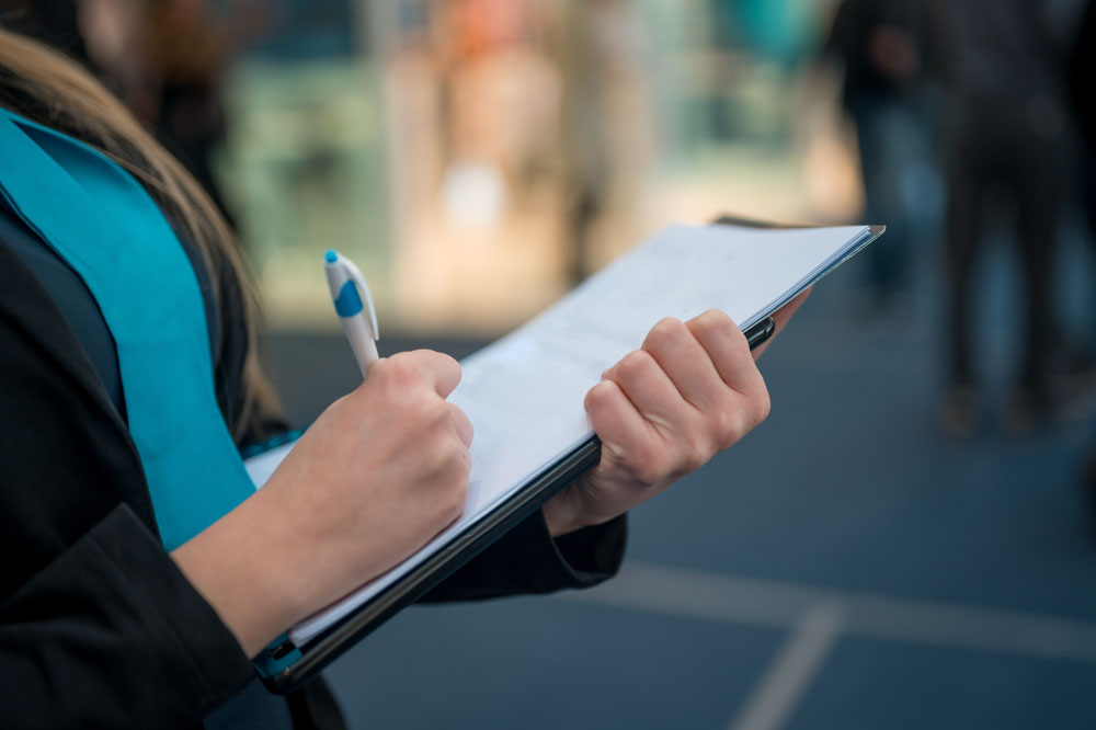 Person in a blue shirt writing with a pen on paper supported by a clipboard